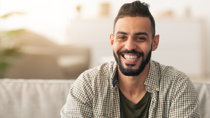 Fototapeta premium A man with a beard sits on a couch, smiling warmly. The living room has soft lighting and a relaxed atmosphere. He appears cheerful and comfortable in his surroundings.