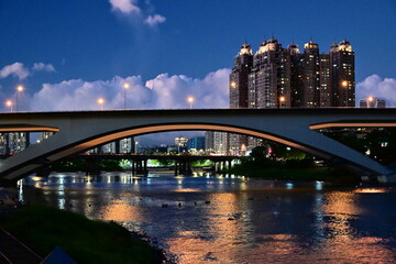 Illuminated arched highway bridge spanning Xindian River at Bitan Scenic Area with reflections on calm water, high-rise buildings glowing against deep blue twilight sky in New Taipei City.