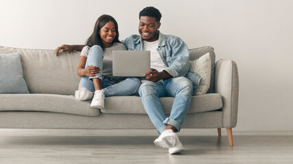Happy loving young African American couple using pc, watching video, looking at their photos or choosing what to buy, sitting and relaxing on couch in living room. Guy embracing lady, free copy space