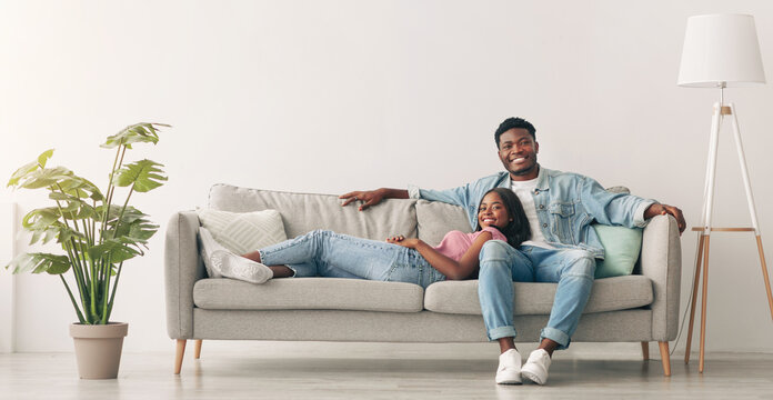 Weekend. Beautiful African American woman and handsome man relaxing on couch at home in living room, smiling couple looking at camera, posing, spending leisure time together, lady lying on guy's lap