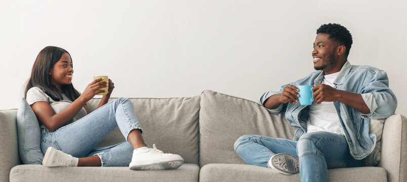 Stay At Home Date. Beautiful African American couple drinking coffee and talking, resting on the couch indoors at home in living room, relaxing enjoying company of each other spending weekend together - Powered by Adobe