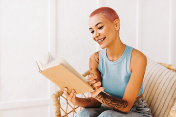 A young woman with a pink shaved head sits comfortably in a chair, reading a book. She appears...