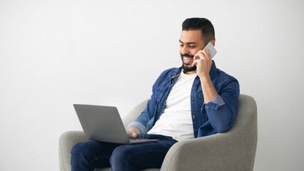 A young man is comfortably seated in a chair, smiling while speaking on his smartphone. He is multitasking by working on his laptop, creating a relaxed home office atmosphere.