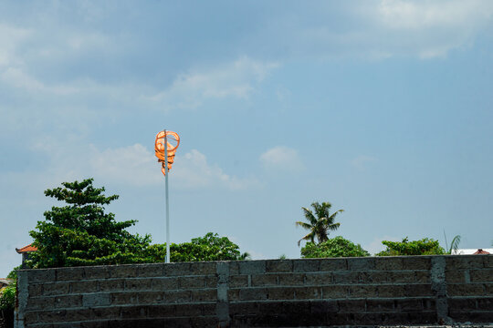 orange windbag on a pole by the sea with a background of green trees with beach umbrellas and small boats around it with a clear sky background for advertising space.