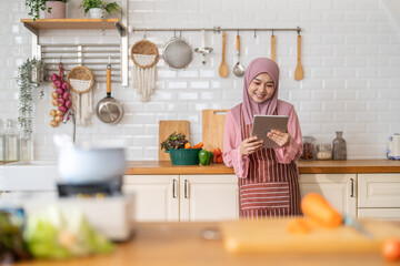 Smiling muslim woman wearing hijab use digital tablet in kitchen while preparing healthy food, online recipe search, home cooking and technology, halal cooking content, food blogger, healthy lifestyle