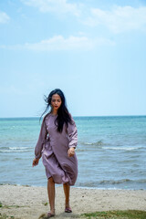 . A long-haired Asian woman wearing a light purple dress with long sleeves walks on the beach with a clear sky background for advertising space.