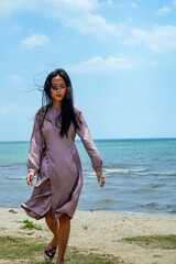 . A long-haired Asian woman wearing a light purple dress with long sleeves walks on the beach with a clear sky background for advertising space.