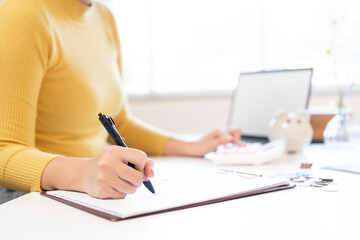 A woman wearing a yellow sweater is writing notes, calculating finances, or managing personal budgets with coins and a calculator on her desk