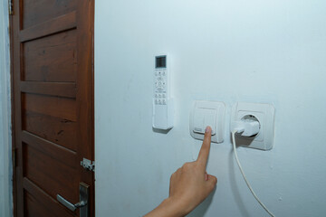 hand trying to turn on the light switch on the white wall with the AC remote control on the left and the socket with the charger cable connected to the right.