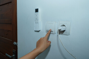 hand trying to turn on the light switch on the white wall with the AC remote control on the left and the socket with the charger cable connected to the right.