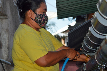 From a low angle, a mother cleans the plastic pipe water drinker in the rear side of the chicken coop using a water sprayer hose. © agratitudesign