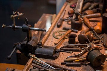 A close-up shot of an antique wooden workbench displays an assortment of metal and wood crafting tools, including a vise and various precision instruments.