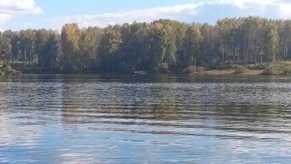 The shaky surface of a beautiful lake surrounded by a dense mixed forest under a cloudy sky on a sunny day in early autumn.