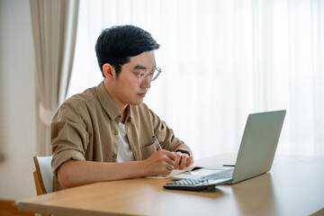 Happy young Asian man working on laptop in living room. Freelance worker male sitting at desk writing notes while watching webinar, studying online