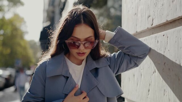 Stylish young woman with sunglasses walking confidently on a busy city street, touching her hair and enjoying the sunlight. Fashionable Young Woman in Sunglasses Enjoying Sunny City Day