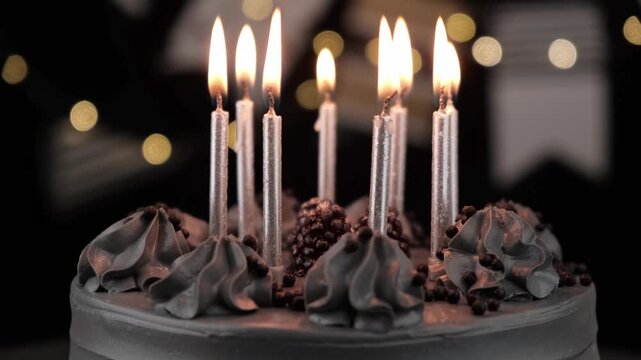majestic black birthday cake with beautifully decorated cream and lit silver candles slowly turning against dark background with festive golden bokeh lights, celebrating special occasion. close up.