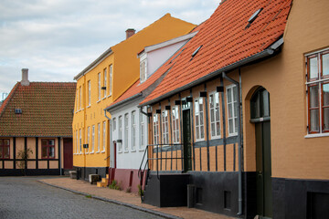 A charming cobbled street is lined with colorful historic houses, each featuring unique architectural details and a traditional aesthetic under a soft, overcast sky.