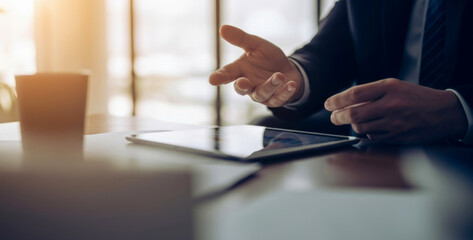 Businessman gestures over tablet device during indoor meeting with warm light, expressing confidence and teamwork as colleagues collaborate around table in bright professional office atmosphere