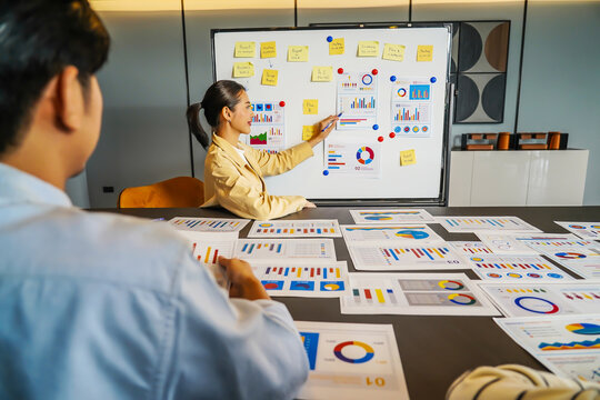 In a vibrant corporate setting, a dynamic young asian businesswoman presents important statistical insights to her engaged colleagues, analyzing data presented on a whiteboard in the office