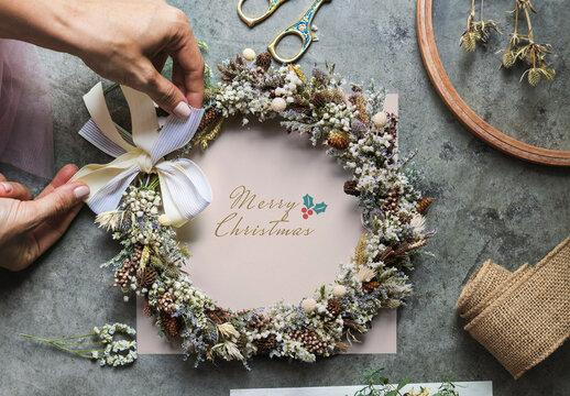 Woman Tying A Ribbon On A Floral Wreath Over A White Card Mockup