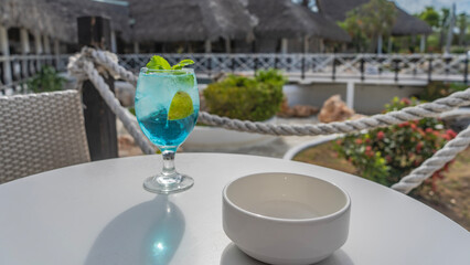 Cocktail on the table in an outdoor cafe. Blue alcoholic drink with ice, mint leaves. Rope railings of the terrace. The soft background is a tropical park, buildings with thatched roofs. Cuba.Varadero