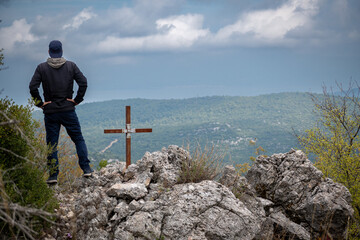 Photo of a man wearing cap and hoodies on a rock next to an iron cross watching the view in the mountains.