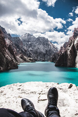 First-person perspective of a person's boots resting on a cliff edge overlooking the vibrant turquoise Kol Suu Lake and mountain canyon