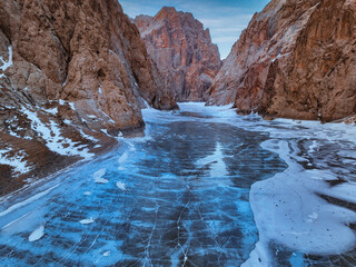 Close-up view of the cracked, frozen surface of Kel Suu Lake leading into a canyon with towering red-brown rock walls