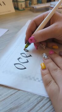 Close-up of female artist hand practicing decorative calligraphy with an ink pen on white paper. Creative lettering process focused on handwriting precision and artistic skill.