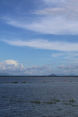 A serene view of a body of water, possibly a lake or river, with clear skies above and clouds scattered in the distance. The water's surface is calm, reflecting the blue sky