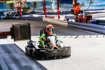 Racer kid wearing helmet and green suit driving a black go kart on an outdoor track, having fun and...