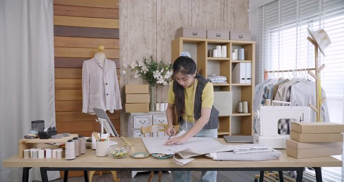 Asian female fashion designer preparing fabric and marking pattern details for handmade clothing inside tailoring studio during small business production workflow with modern tools