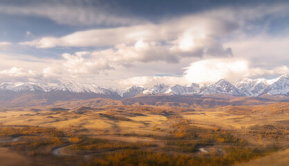 Colorful autumn landscape in the Altai region features rolling hills, golden trees, and majestic snow-covered mountains under a dramatic sky during daylight.