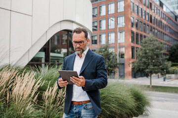 Fototapeta premium Businessman in front of office building, holding tablet and reading something.