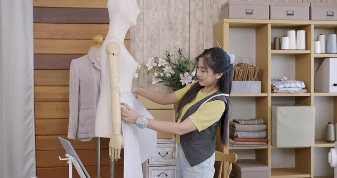 Asian female fashion designer preparing paper prototype on mannequin by aligning centerfold and measuring position for handmade clothing creation in tailoring studio workspace environment