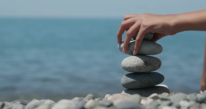 Female hand carefully placing stones on top of each other to create a balanced stack on a pebble beach, with the tranquil blue sea in the background