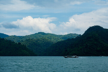 Rocky mountains, lush with trees, rise from the azure water, their cliffs catching the afternoon sun, set against a sky filled with clouds.