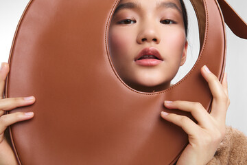Asian woman models a stylish leather round bag in a studio setting, creating a chic fashion moment. Her poised expression and close framing emphasize texture, color, and contemporary style.