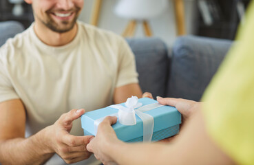Close-up cropped photo of female surprises her partner with small gift box in cozy, modern living space, hands him present, congratulate with life event, birthday, special occasions or anniversary