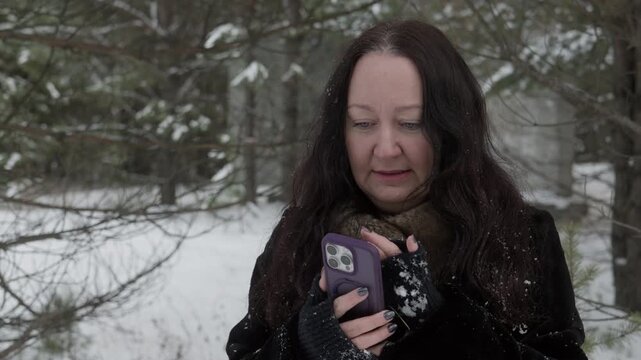 Middle-aged woman with dark hair wearing black coat and brown fur collar holding purple smartphone case, checking phone in winter forest. Gentle smile, natural light.