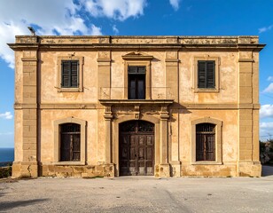 An old, weathered beige building with dark shutters and doors sits under a blue sky with fluffy clouds. The structure is symmetrical