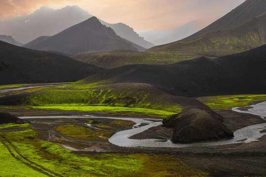 A winding river cuts black volcanic hills and moss plains near Landmannalaugar, Iceland, at dawn or sunset, warm light and mist softening layered mountains.