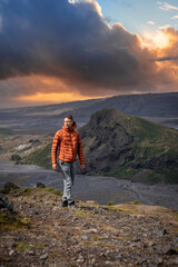 Naklejka premium A lone hiker walks a rocky ridge at sunset in Iceland highlands. Storm clouds glow above mossy lava slopes and a gray riverbed near a craggy peak, with rich contrast.