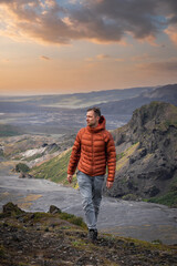 A lone hiker in an orange jacket walks a mossy ridge in Iceland, framed by layered mountains and a basalt outcrop in low evening light with deep depth of field.