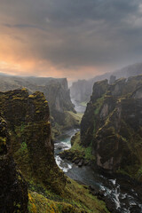 Wide angle view of Fjadrargljufur Canyon in southern Iceland, sheer cliffs and a dark winding river under low clouds and mist, warm sunset glow, long exposure water
