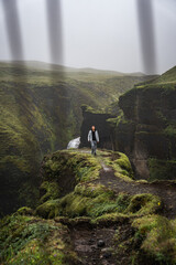 A lone hiker walks a narrow moss ridge above a deep canyon in Iceland as rain falls and mist...