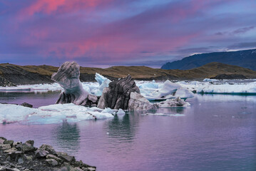 Blue white icebergs streaked with ash rest in a calm lagoon at Jokulsarlon, Iceland. Wide framing and soft light at twilight reflect pastel tones and distant dark mountains.