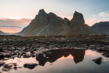 Jagged Vestrahorn rises over black sand and pebbles at Stokksnes, Iceland, twin peaks reflecting in a calm tidal puddle at sunset with soft pastel light.