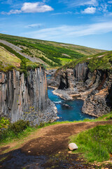 Midday summer light defines Studlagil Canyon in East Iceland as a turquoise river runs between hexagonal basalt columns, with green hills and a trail in the foreground.