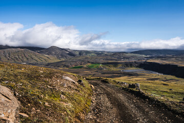 A gravel track winds over a mossy volcanic plateau in Iceland, leading toward basaltic mountains with low clouds. Bright daylight and long shadows reveal rugged lava textures.